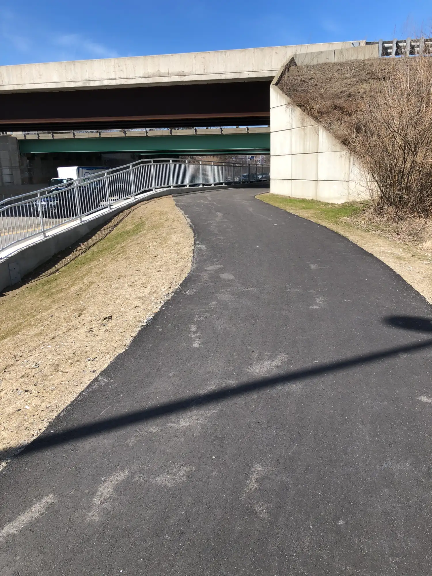 WESTBROOK STREET SHARED USE PATH 3 (1) - Sebago Technics A paved pedestrian path curves uphill toward an overpass, bordered by grass and a metal fence on a sunny day.