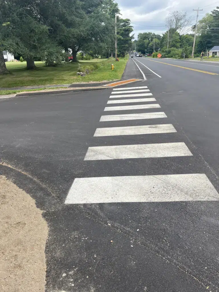 WINDHAM ACTIVE TRANSPORTATION PLAN 2 - Sebago Technics A crosswalk with white stripes leads across a newly paved road in a suburban area with trees and grassy lawns on the roadside.