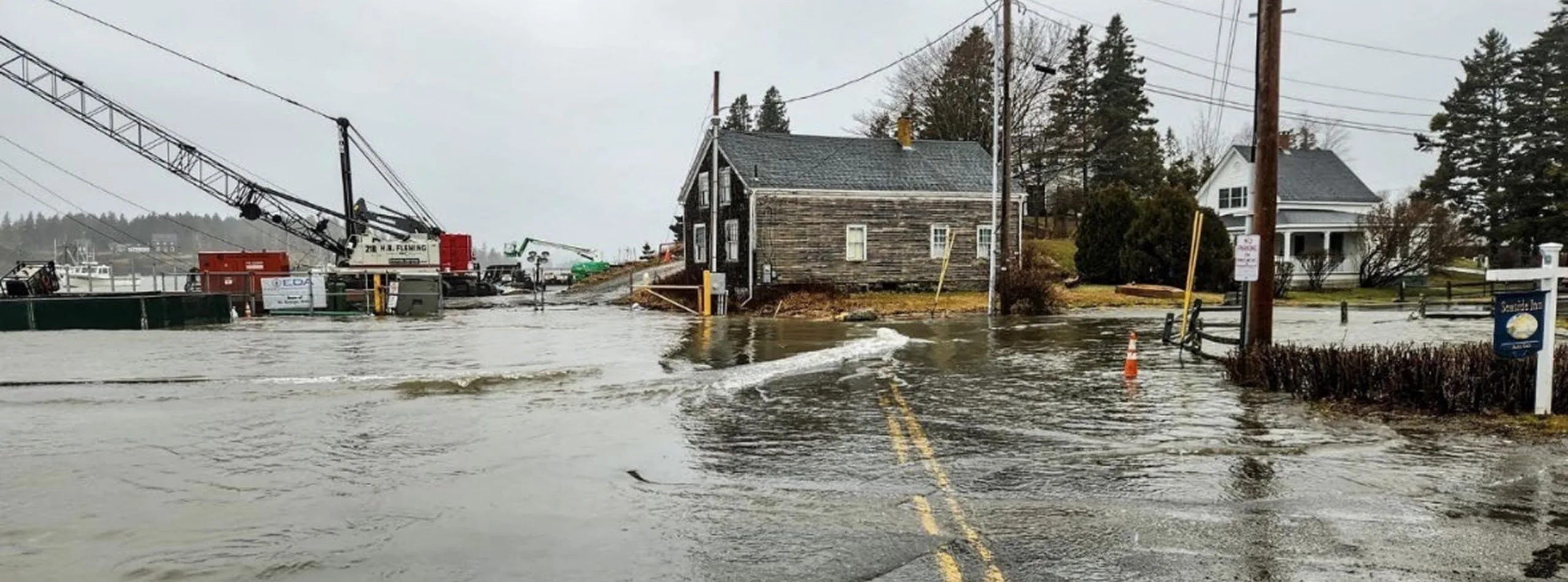 Flooded street with water covering the road, houses on either side, and a Maine engineering firm’s construction equipment in the background under a cloudy gray sky.