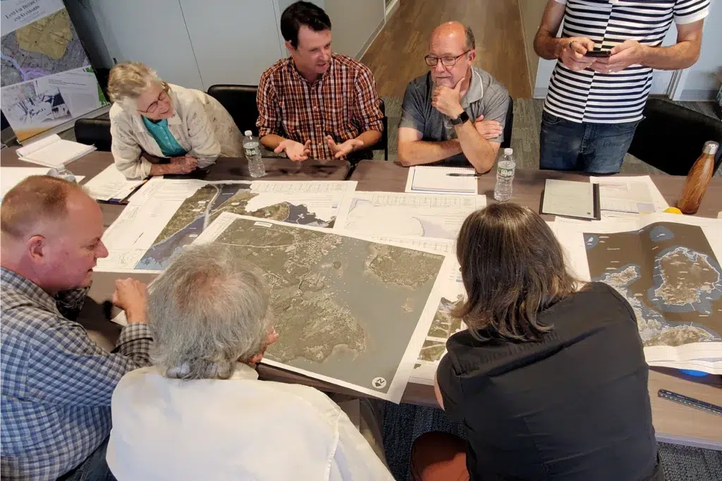 st-george-our-approach - Sebago Technics Six people sit around a table covered with maps, discussing and pointing at details, suggesting a Maine engineering firm’s group meeting or planning session.